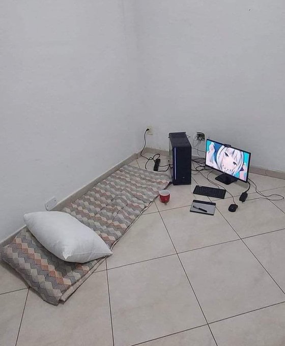 A minimalist setup on a tiled floor with a mattress, pillow, computer tower, monitor displaying an anime character, keyboard, mouse, and a small red cup.