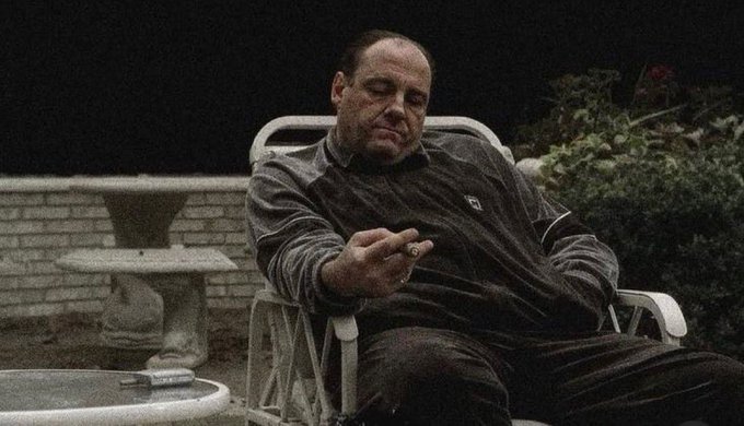 An older man in a dark tracksuit sitting in a patio chair, looking down while holding a cigar, with a stone fountain and greenery in the background.