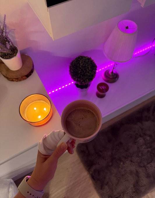 A first-person shot of a hand holding a creamy latte, paired with a "gm" crypto trader greeting wishing big profits to start the day. The cozy candle-lit setup with purple LED lighting captures the relaxed morning ritual vibe of checking charts with coffee in hand.