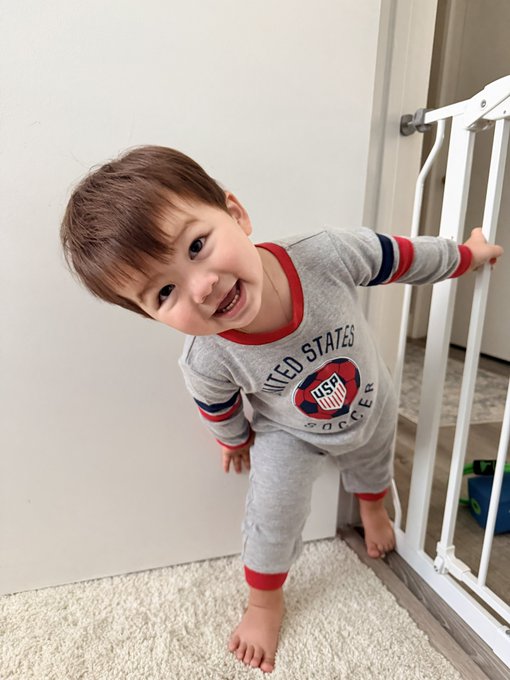 A young child with brown hair smiles cheerfully while standing on a carpet near a baby gate, wearing gray 'United States Soccer' pajamas with red and blue trim.