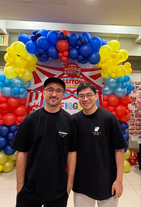 Two smiling men stand in front of a balloon arch at a Jollibee Kids Party event, with a banner reading 'FICC' partially visible in the background.