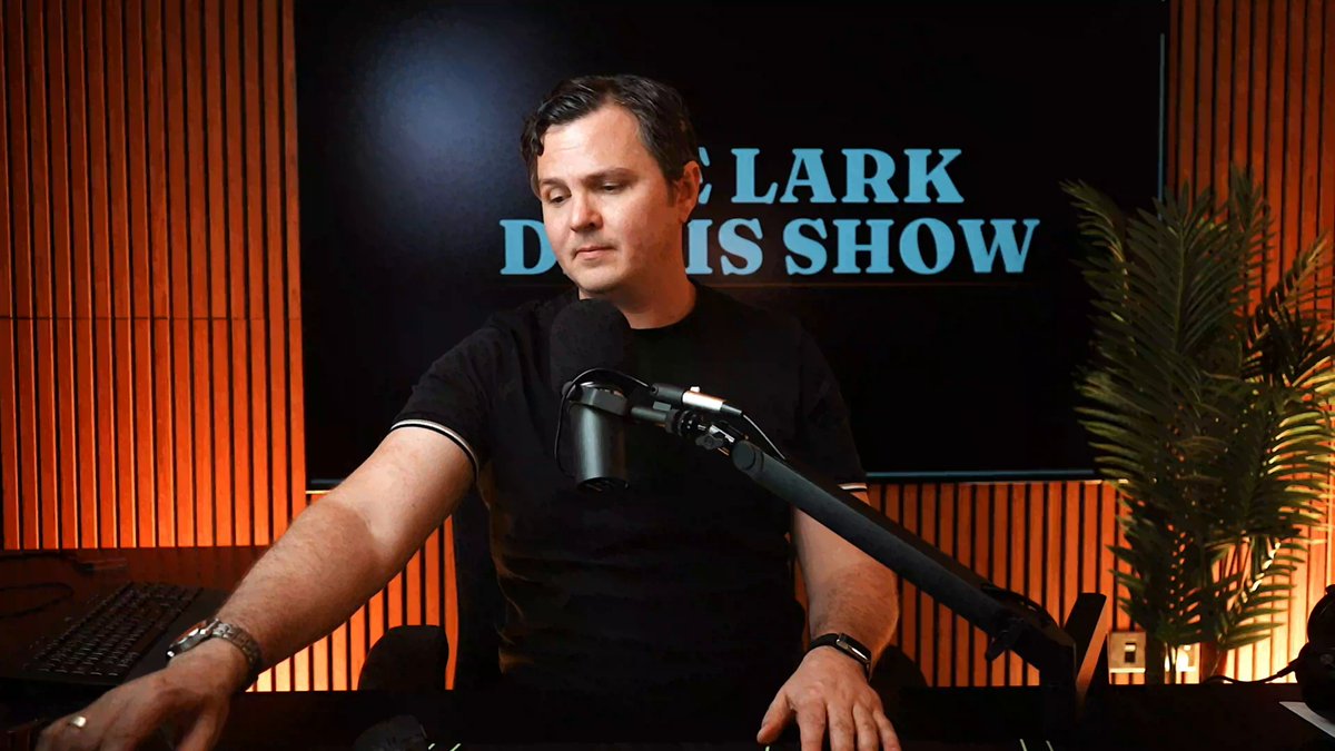 The image shows a man sitting at an office desk with a microphone, possibly recording something or giving a live broadcast. Behind him, there is a large screen displaying text related to Bitcoin and the stock market updates, such as Bitcoin just flipped 0:00 and Ethereum also just flipped 1:40. There are charts showing Bitcoin and Ethereum fluctuations over time as well. The room has wood paneling on the walls, creating an office atmosphere.