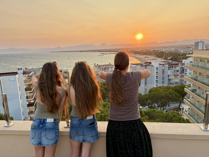 Three young women with long hair stand on a balcony overlooking a coastal town and ocean at sunset, with the sun setting behind distant mountains.