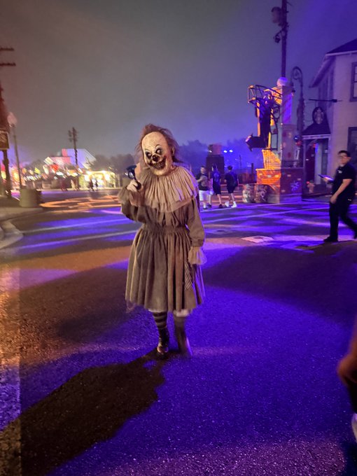 A person in a creepy clown costume stands on a street illuminated by purple and orange lights, likely at a Halloween horror event.