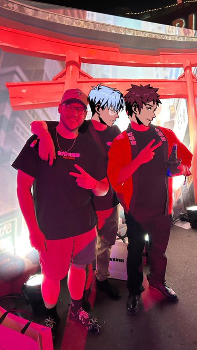 A man wearing a "STREETS OF AZUKI" t-shirt poses with life-sized cutouts of Azuki NFT anime characters in front of a red torii gate at what appears to be a Street of Azuki event. The setup reflects his dedication to the Azuki collection, with a placard labeled "ARUKI" visible in the background, tying the fan moment to the brand's immersive experience.
