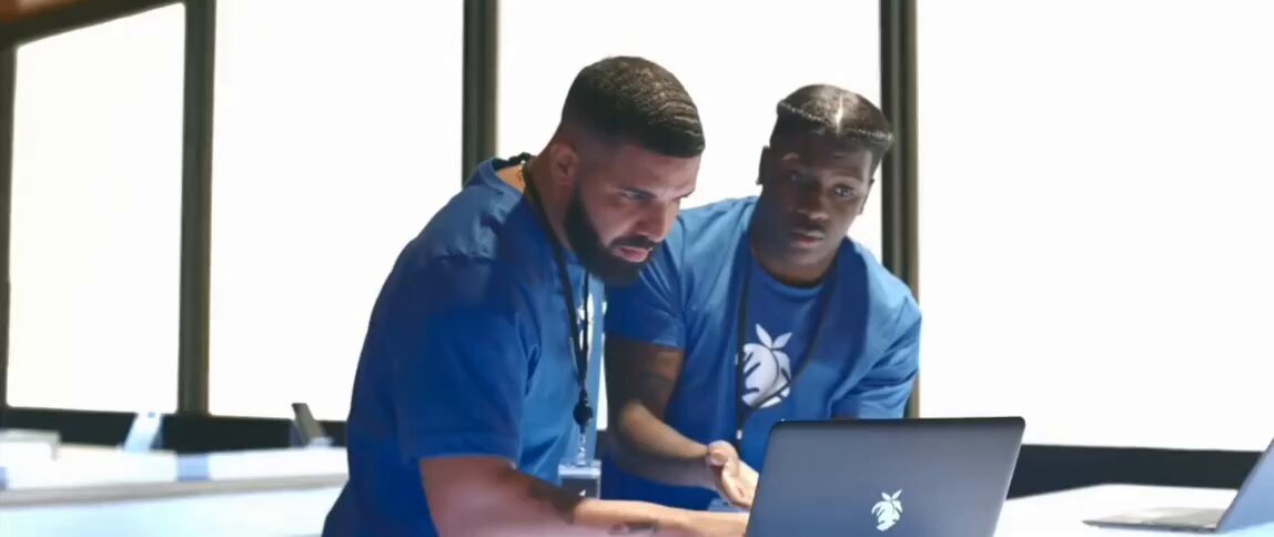 The image shows two men sitting at a table with laptops open in front of them. They appear to be discussing something, possibly related to their work or research. The background features a window that allows natural light to enter the room, creating a comfortable atmosphere for their conversation.