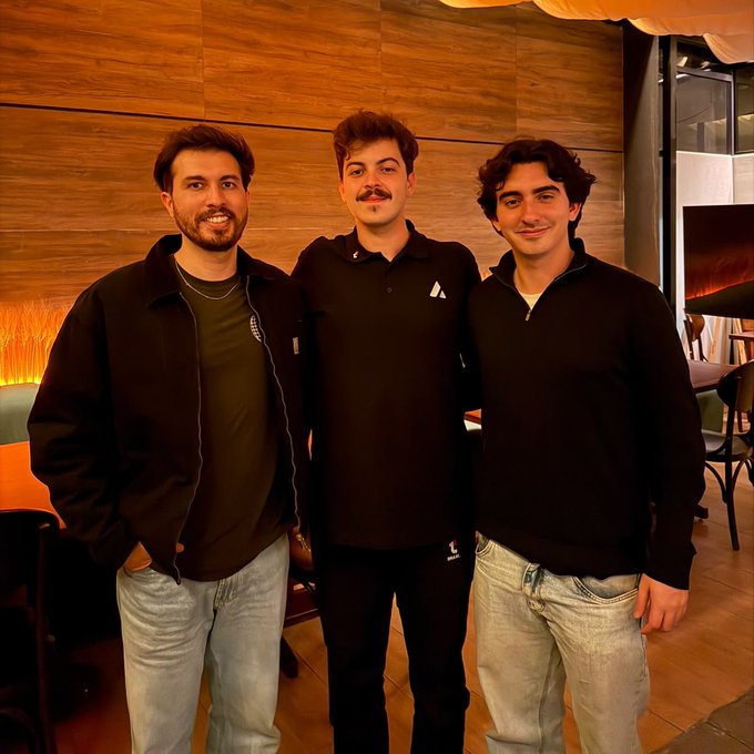 Three young men pose together indoors, wearing dark tops and jeans, in front of a wood-paneled wall.