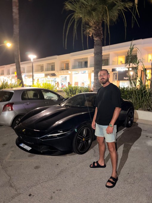 A man in a black t-shirt, denim shorts, and slides stands proudly next to a sleek black Ferrari parked outside a row of lit-up shops, palm trees towering in the background at night—perfectly matching a casual "Gm friends" vibe with a touch of luxury.