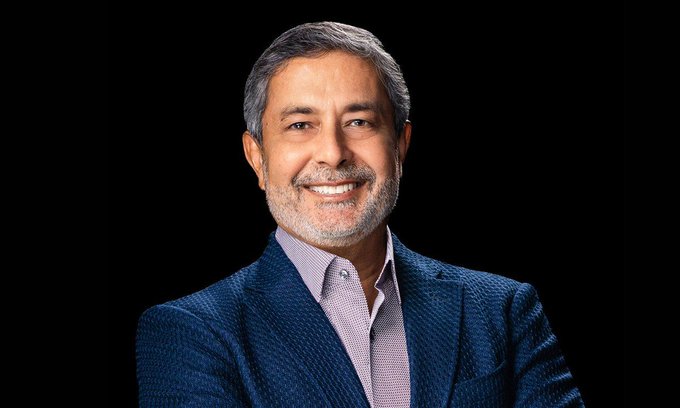 The image features a man wearing a blue suit jacket, smiling at the camera with his arms crossed over his chest. He has gray hair and a beard, giving him an older appearance. The background of the photo is black, which contrasts with the man's attire and makes him stand out more. There are no other objects or texts in the image, so it focuses solely on the man and his expression.