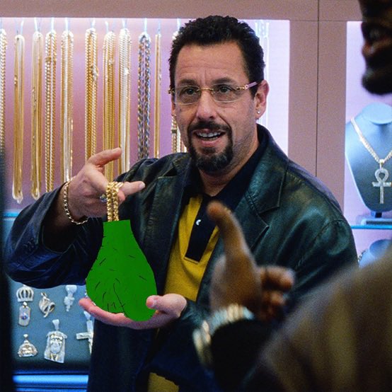 Adam Sandler in a jewelry store scene (from Uncut Gems) holding a large green pendant bag-shaped object covered in exclamation marks while pointing at it.