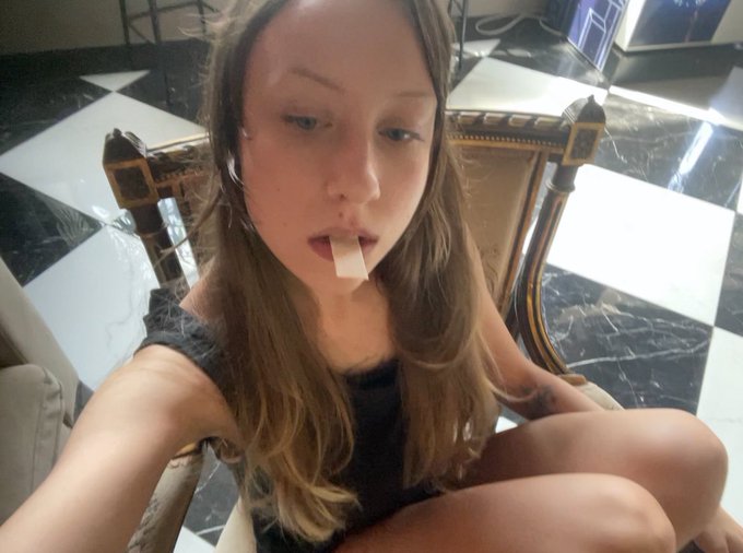 A young woman with blonde hair sits in an ornate chair on a checkered floor with a piece of tape over her mouth.