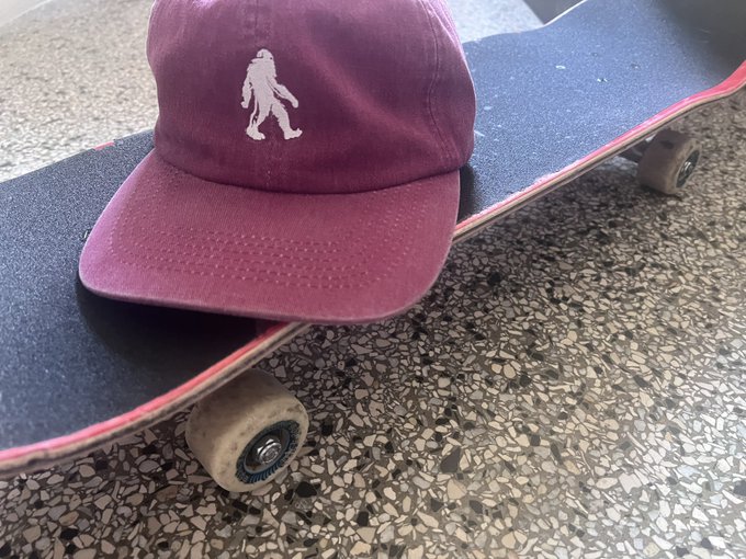 A faded maroon baseball cap featuring a Bigfoot/Sasquatch silhouette logo resting on top of a skateboard on a speckled floor.