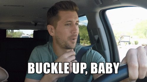 The image shows a man wearing a green shirt, sitting inside a car with his hand on the steering wheel. He appears to be driving or preparing to drive. There are two other people visible in the background of the image, but they do not seem to be involved in any activity related to the main subject. The text "Buckle up, baby" is written above and below the man's head, which could possibly be a caption for the image or part of an advertisement.