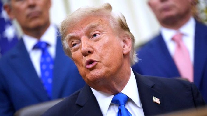 Donald Trump, wearing a dark suit and blue tie, looks off to the side while speaking, with other suited figures visible in the background.