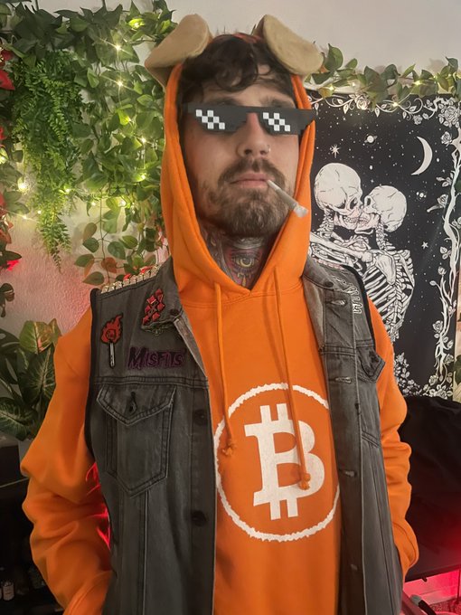 A person wearing an orange Bitcoin hoodie, denim vest with patches, pixelated sunglasses, and a novelty cigarette, stands in front of a skeleton tapestry.