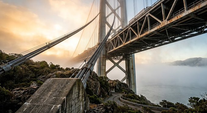 A dramatic view of the Golden Gate Bridge, showing its massive steel structure, suspension cables, and a tower emerging from low-lying fog above a rocky coastline.