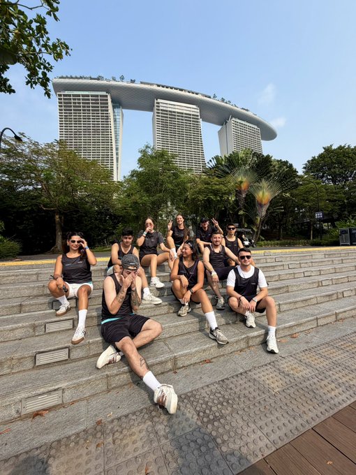 A group of athletes in matching athletic gear poses on stone steps with the iconic Marina Bay Sands hotel towering in the background after a morning run, sponsored by Fhenix as part of a Token2049 event.