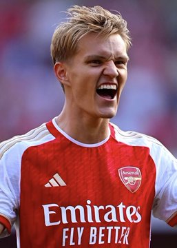 A blonde Arsenal player in a red and white home jersey featuring the "Emirates FLY BETTER" sponsor logo and the club crest yells passionately during a match, likely representing the team's effort behind the bold $3.1M wager placed on them to defeat Aston Villa.