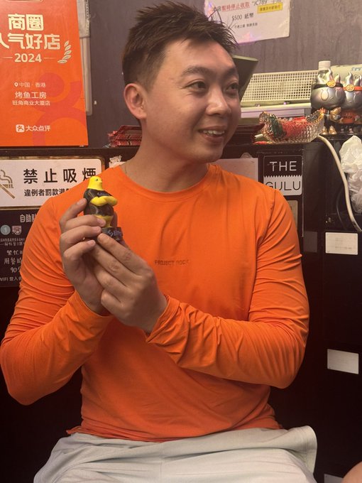 A man wearing an orange long-sleeve shirt smiles while holding a small figurine (appearing to be a character with a yellow hat), likely one of the "奶印" collectibles referenced in the post. Behind him, posters display Chinese text including "商家人气好店 2024," "禁止吸烟" (No Smoking), and a sign reading "THE GULU," suggesting the photo was taken in a popular, award-recognized shop or restaurant setting.