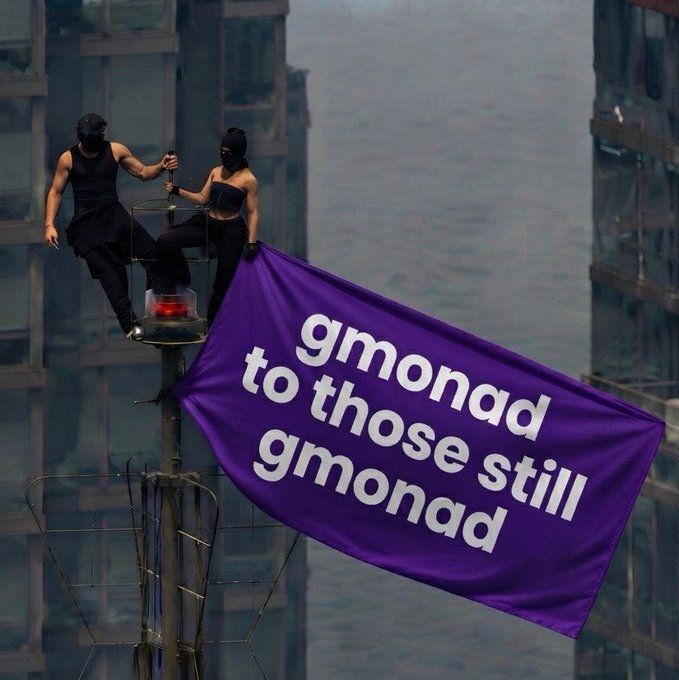 Two masked performers in black stand on a tall structure displaying a purple banner that reads 'gmonad to those still gmonad'.