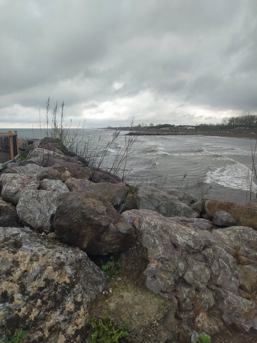 A rocky shoreline overlooks a turbulent, grey ocean under a heavily overcast sky, with a distant coastline visible on the horizon.
