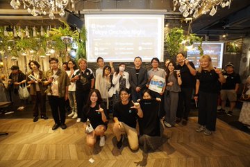 The image shows a group of around fifteen attendees posing together at what appears to be a FomoWell event, with a presentation screen in the background displaying "FomoWell Tokyo Ecosystem Night" along with a date and "Powered by Bitget Wallet" branding. The lively group is gathered in a stylish venue featuring elegant chandeliers and greenery, capturing the successful conclusion of the Tokyo gathering.