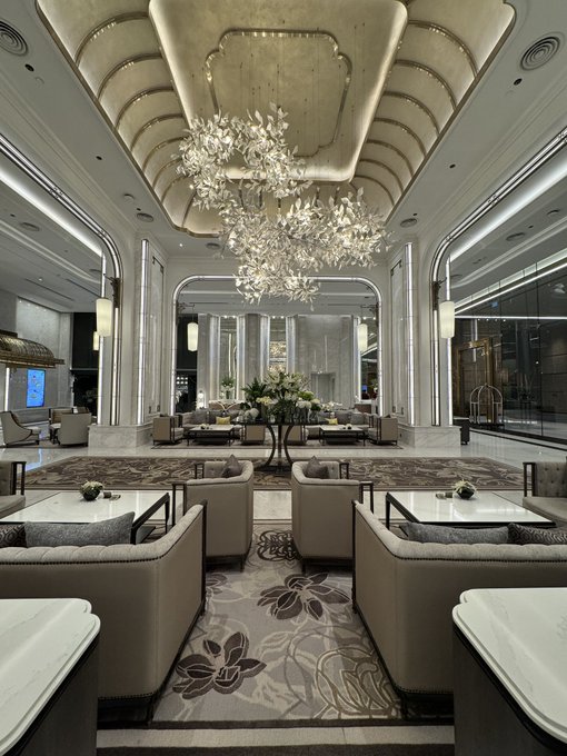 An elegant hotel lobby or lounge with tall columns, plush seating, marble tables, patterned carpet, and ornate crystal chandeliers hanging from a decorative ceiling.