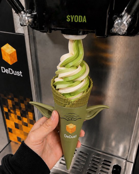 The image features a person holding an ice cream cone with green and white colors, which appears to be a creative design inspired by the Star Wars character Yoda. The ice cream cone has a unique shape resembling a Yoda-like figure or a robot, adding a fun and playful twist to the traditional ice cream experience.

The image also includes text that reads "Creative the TON community is... impressed Yoda is". This suggests that the person holding the ice cream cone might have received positive feedback from others for their creative design choice of an ice cream cone inspired by Star Wars, which could be a source of pride and satisfaction.

The image also includes a reference to "DeDust" as a brand or product, indicating that the ice cream cone is likely made by this company.