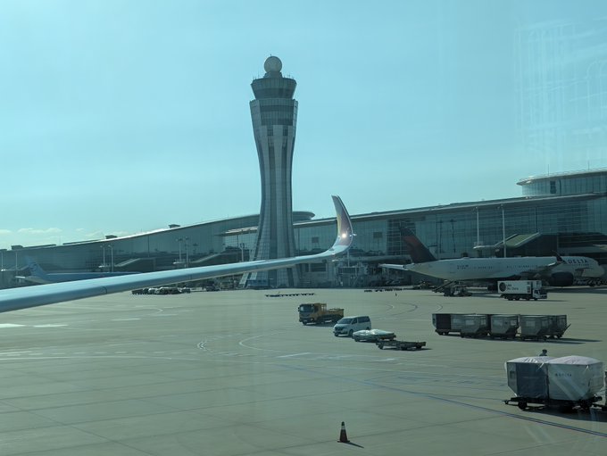 A view from an airplane window showing an airport tarmac with a tall air traffic control tower, parked planes including a Delta aircraft, ground service vehicles, and a terminal building under a hazy sky.