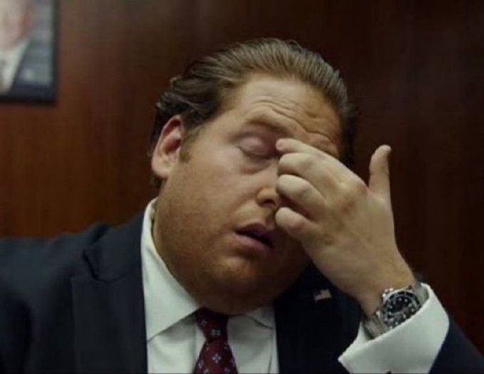 A man in a dark suit, white shirt, and patterned tie rubs his face and eyes with his hand, wearing a silver wristwatch, appearing tired or frustrated.