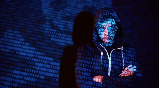 A hooded figure in a dark hoodie illuminated by blue binary code, with one hand raised, set against a backdrop of projected code and shadow.