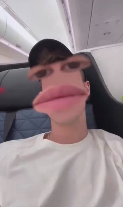 A selfie of a young man wearing a black cap and white t-shirt sitting in an airplane seat, distorted by a humorous filter that enlarges his lips and eyebrows.