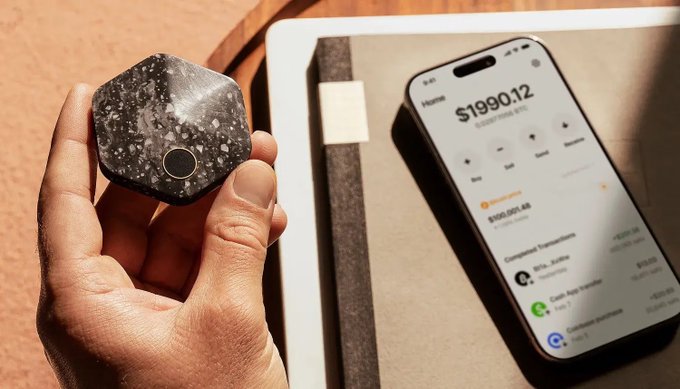 A hand holds a small black octagonal device next to a smartphone displaying a balance of '$1990.12' with various transaction details.