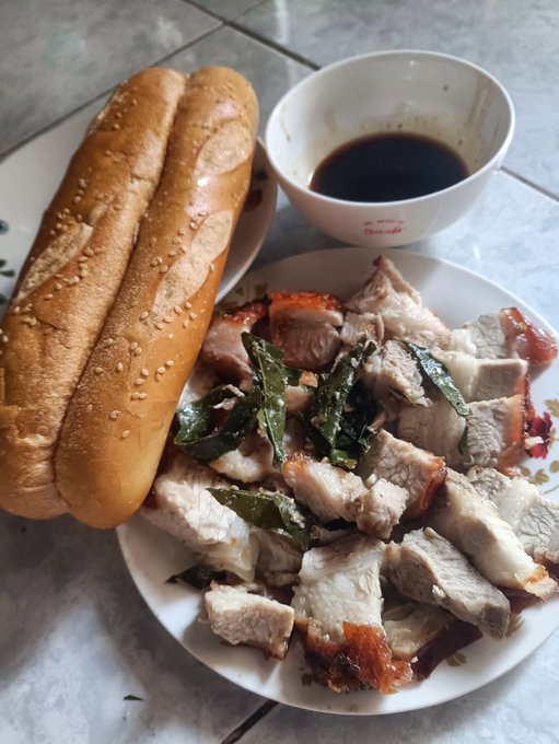 A plate of sliced roasted pork belly topped with fried herbs, served alongside French bread rolls and a small bowl of dark dipping sauce.