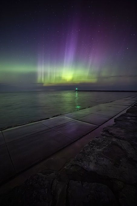 A nighttime landscape photograph capturing the vibrant green and purple aurora borealis stretching across a starry sky above a dark body of water and a stone breakwater, with a small green navigation light reflecting on the water.