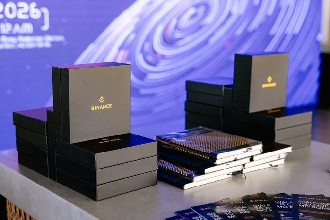 Black product boxes branded with the gold "BINANCE" logo are displayed on a counter, alongside stacks of notebooks and promotional cards. A digital screen in the background shows text with "12AM" and a galaxy-like graphic.