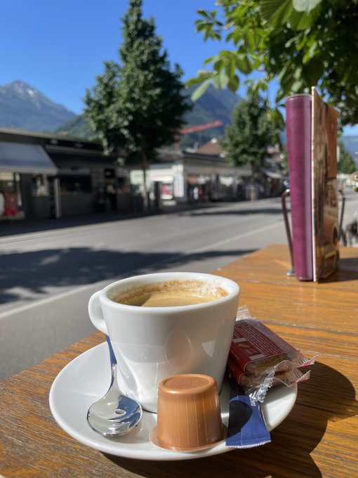 A small espresso cup on a saucer with a chocolate candy and sugar packet sits on a wooden table, with a sunny street, buildings, and mountains in the blurred background.