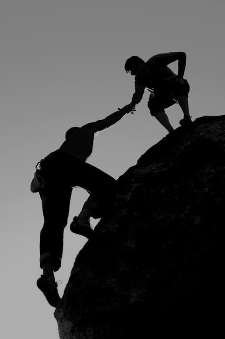 A black and white silhouette photograph of two climbers, with one person reaching down to help pull another climber up a rocky cliff.