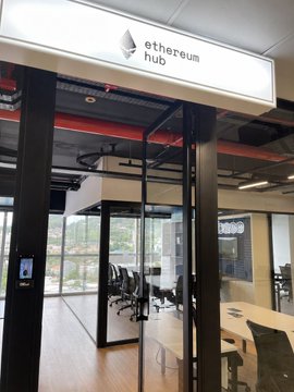 An entrance sign reads 'ethereum hub' above glass doors leading into a modern office space with visible desks, chairs, and exposed ceiling pipes.