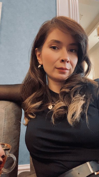 A selfie-style photograph of a woman with long, wavy brown hair with blonde highlights, wearing a black top and earrings, sitting indoors with a blue wall behind her.