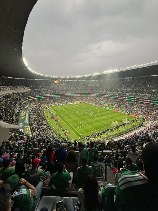 A wide shot of a packed soccer stadium at dusk with fans predominantly wearing green (likely Mexico supporters), showing the field below with players and a large crowd filling the stands under an overcast sky.