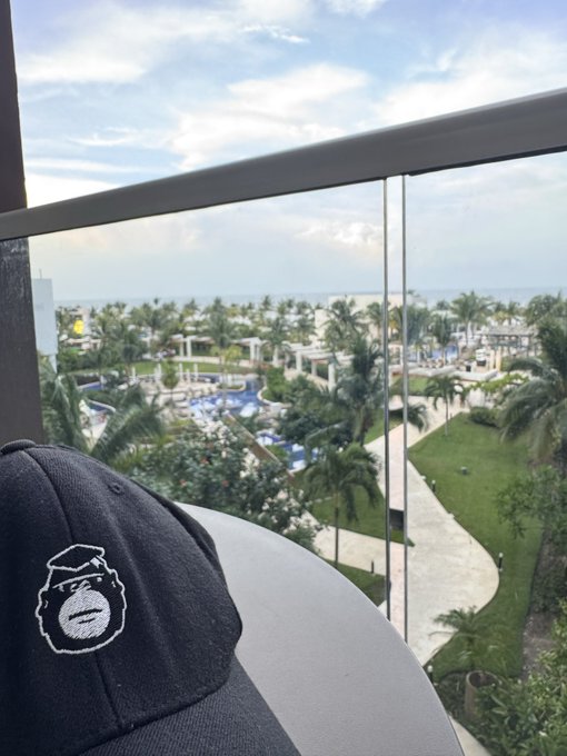 A black baseball cap featuring a white embroidered logo rests on a balcony railing, overlooking a tropical resort with palm trees, a pool area, and the ocean in the distance.