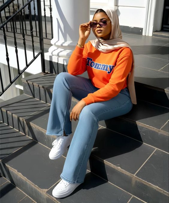 A woman wearing a hijab, orange "Tommy" sweatshirt, blue jeans, and sunglasses sits on black outdoor steps adjusting her eyewear.