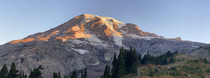 A landscape photograph of a large snow-capped mountain peak at sunrise or sunset, with evergreen trees in the foreground.