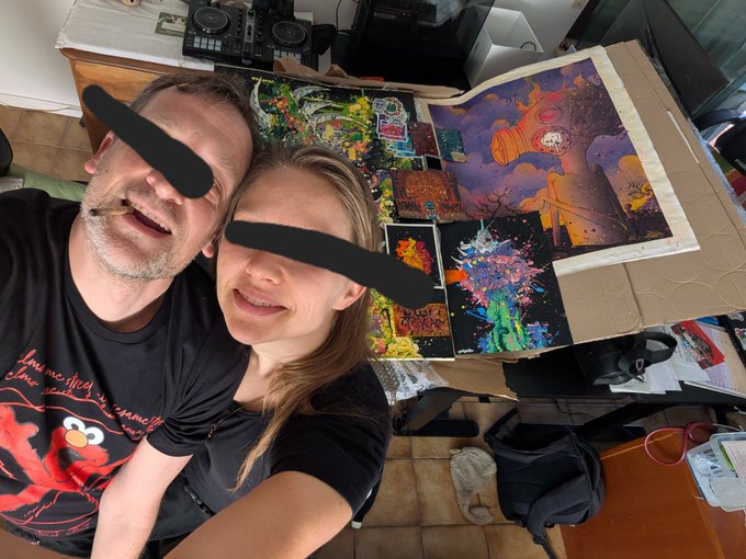 A selfie of two people with black bars over their eyes, taken in a room filled with colorful artwork, comic books, and a DJ controller on a cabinet.