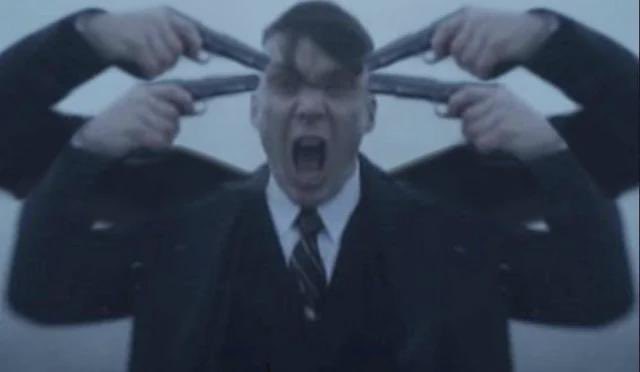 The image features a man dressed in a suit holding multiple guns above his head, with his mouth open as if he's shouting or screaming. The background appears to be a blurred image of another person, possibly indicating that this scene takes place in an indoor setting like a room or studio. The overall composition and the presence of the guns suggest that it could be related to a movie poster for a film featuring a character with a unique appearance and a dramatic backstory involving gun violence.