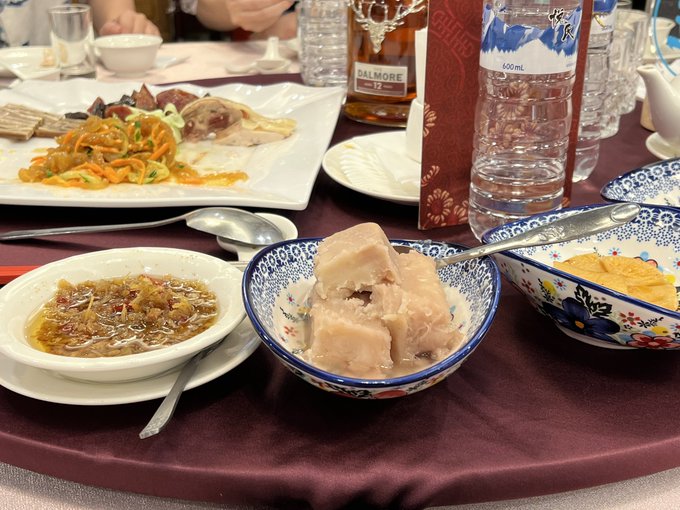 A table set with various dishes including noodles, meat, tofu, and a bowl of soup, alongside a bottle of Dalmore 12 whisky and water bottles on a maroon tablecloth.
