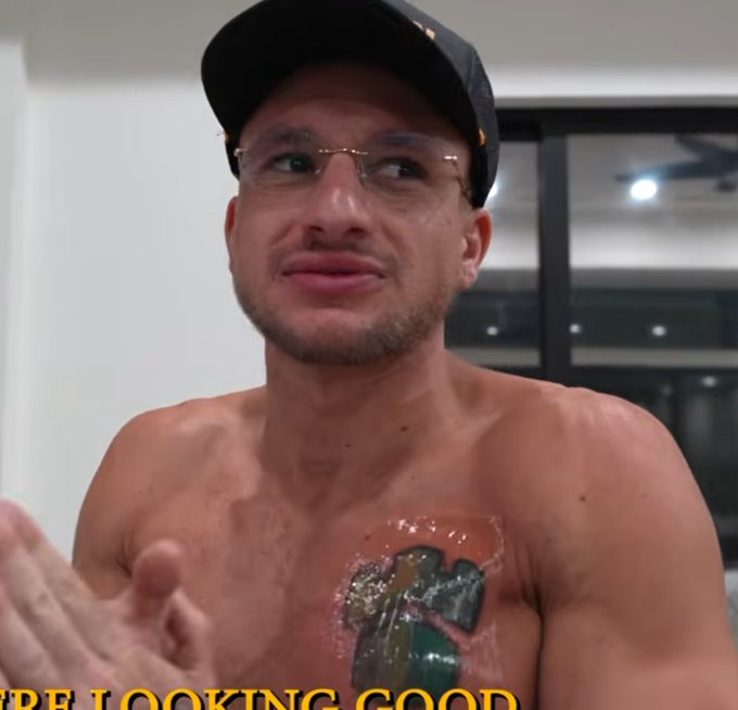 A shirtless man wearing a cap and glasses looks off to the side while gesturing, with a fresh, tattoo of the Gamdom logo prominently displayed on his pectoral. The caption "'RE LOOKING GOOD" is visible at the bottom, and the image illustrates the post's joke about someone named Steve" getting the Gamdom brand permanently inked on his chest.