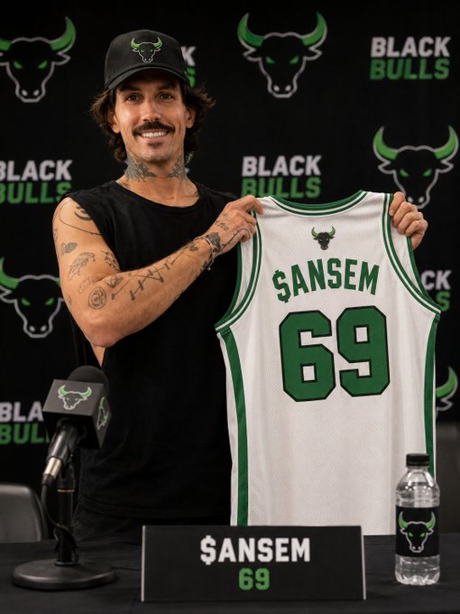 A tattooed man wearing a Black Bulls cap and shirt holds up a jersey with the name "ANSEM" and number 69 at a press conference, set against a Black Bulls backdrop. The image ties to the post's theme of $ANSEM joining the Black Bulls team, as evidenced by the branded water bottle and nameplate on the table—visually reinforcing his announced role as the "unofficial-official Head of Bull Posting" for the meme coin.