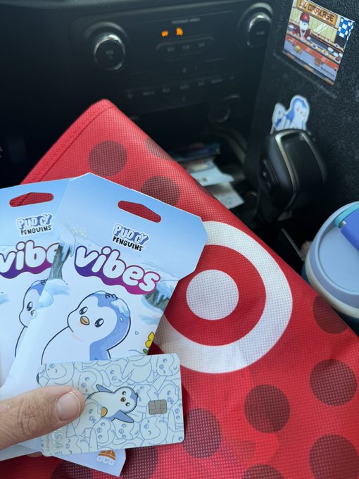 The image shows a hand holding two VIBI VIBES cards, which are small rectangular cards with cartoon penguins on them. The hand is positioned over a red Target shopping bag that has a target design on it. The text of the post reads "Great start to the day. Checked the mail and received my @KASTxyz card. Checked the local @Target and was able to get my hands on some @pudgypenguins @vibes cards".